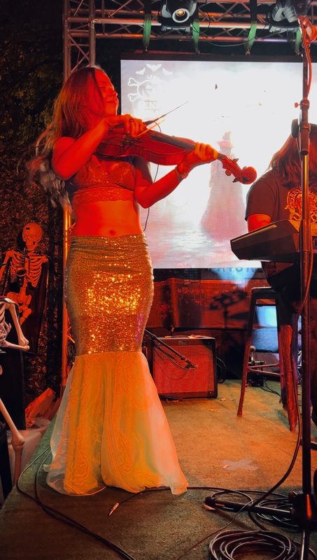 Brianna in a sequined mermaid costume, playing viola on a stage under warm lights